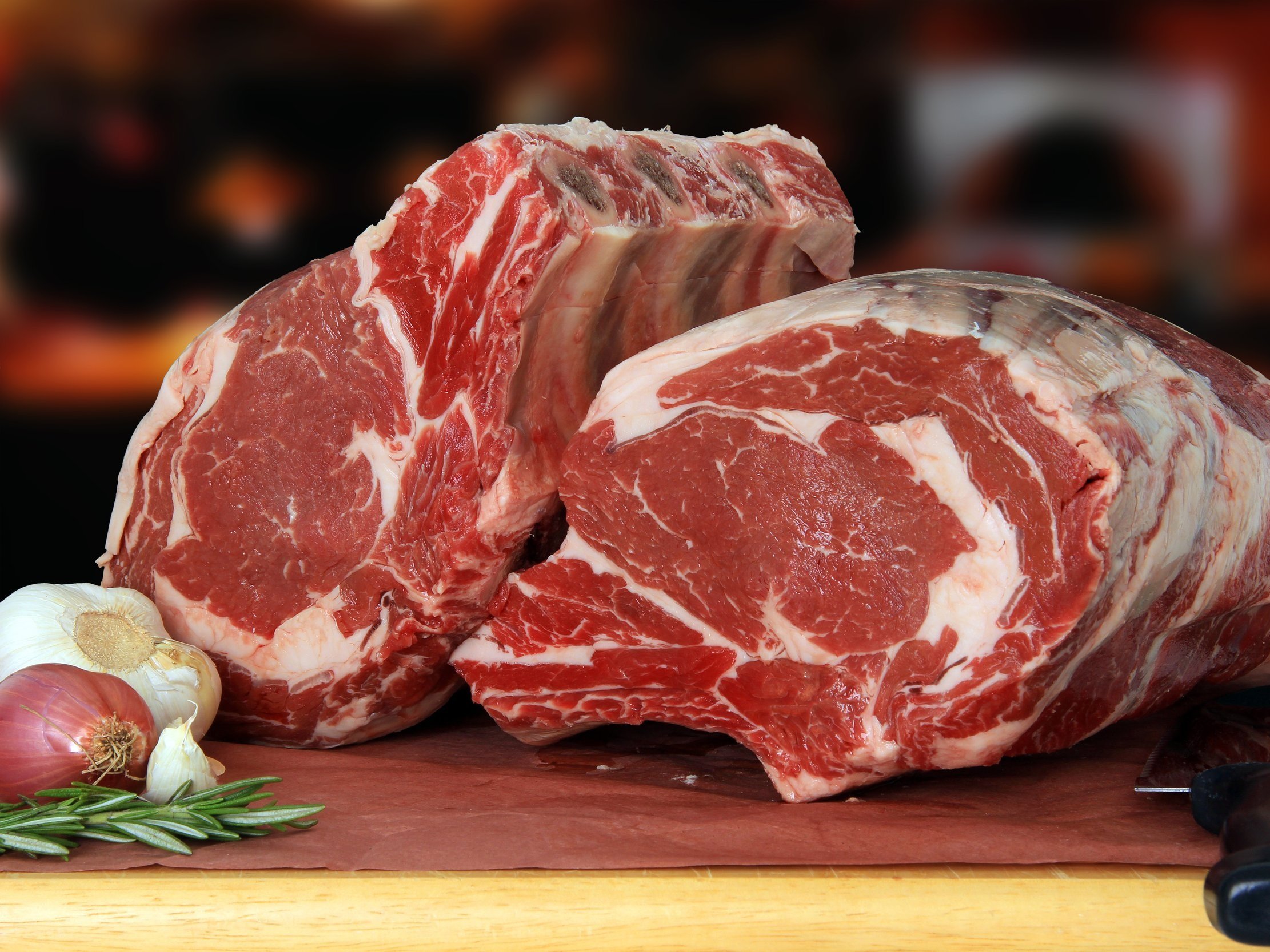 Freshly cut ribeye steaks displayed on butcher paper, accompanied by garlic, shallots, and rosemary, highlighting premium quality meat for culinary use.